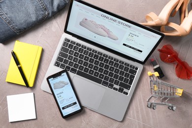 Photo of Online store website on laptop screen. Computer, smartphone, shopping cart, stationery and accessories on grey table, flat lay