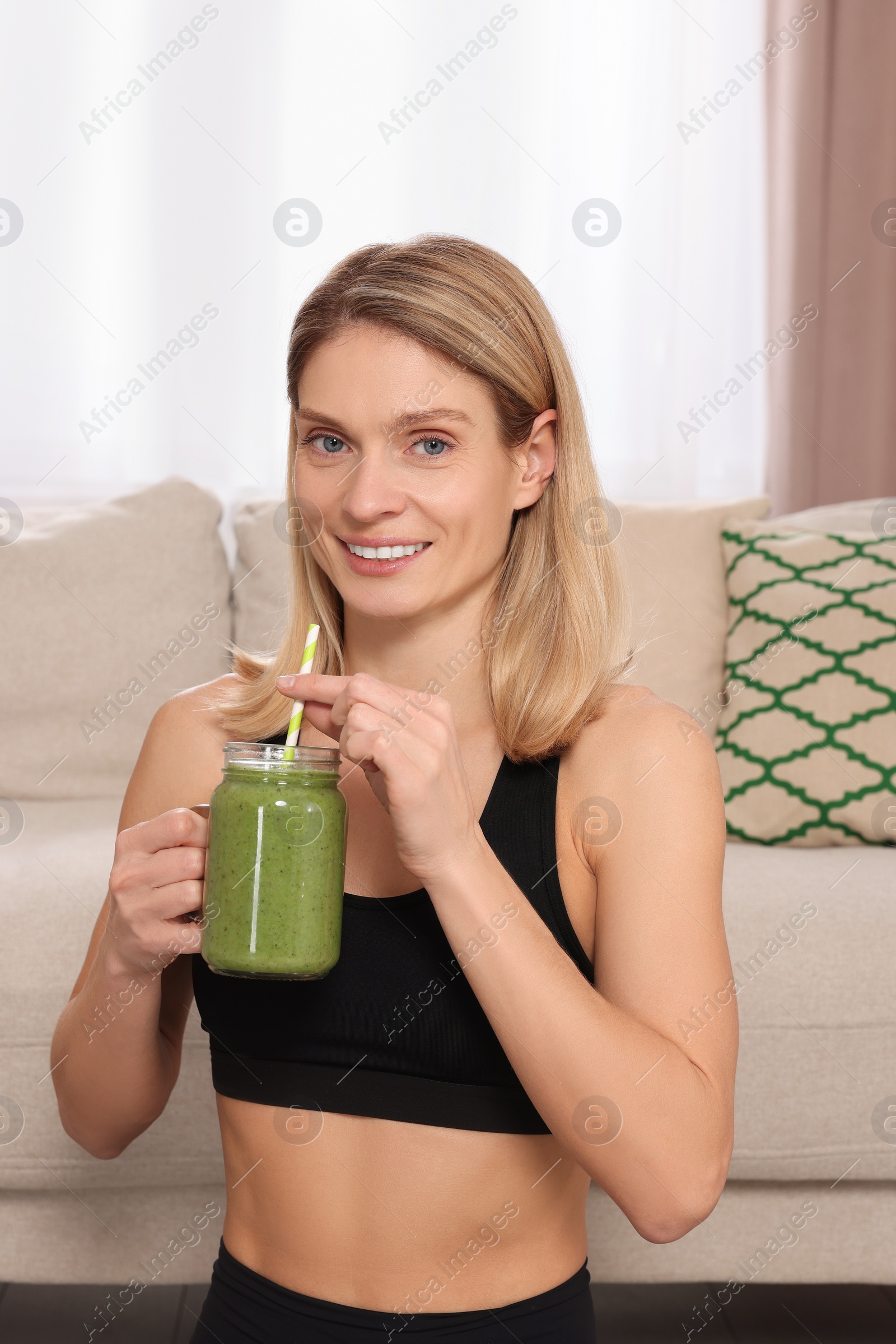 Photo of Young woman in sportswear with mason jar of fresh smoothie at home