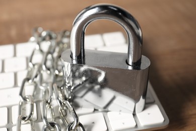 Photo of Cyber security. Keyboard with padlock and chain on wooden table, closeup