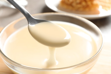 Photo of Spoon of pouring condensed milk over bowl on table, closeup. Dairy products