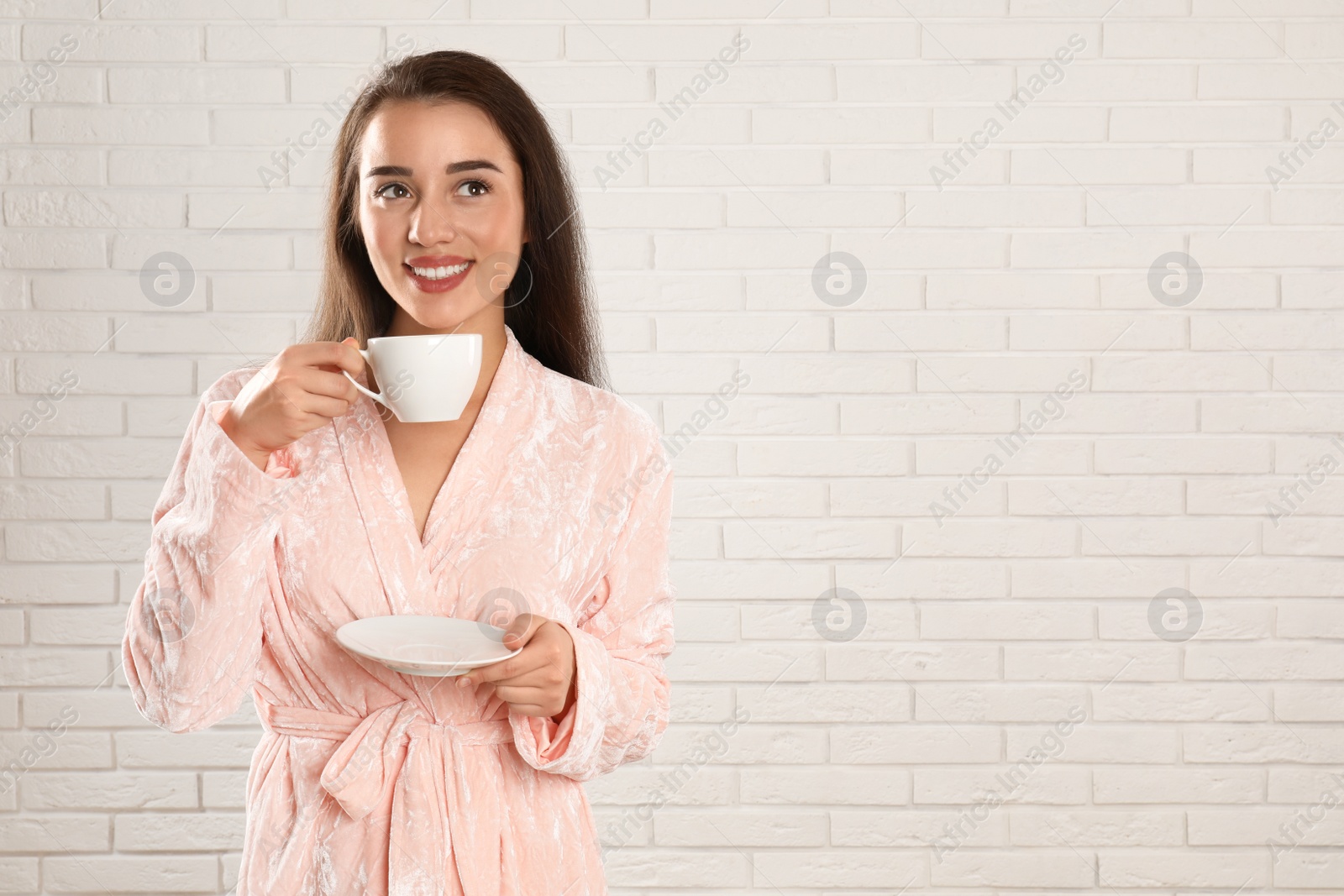 Photo of Young woman in bathrobe with cup of coffee near white brick wall. Space for text