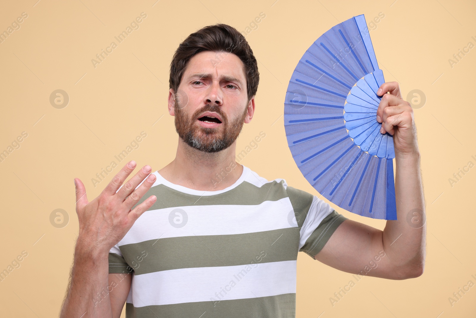 Photo of Unhappy man with hand fan suffering from heat on beige background