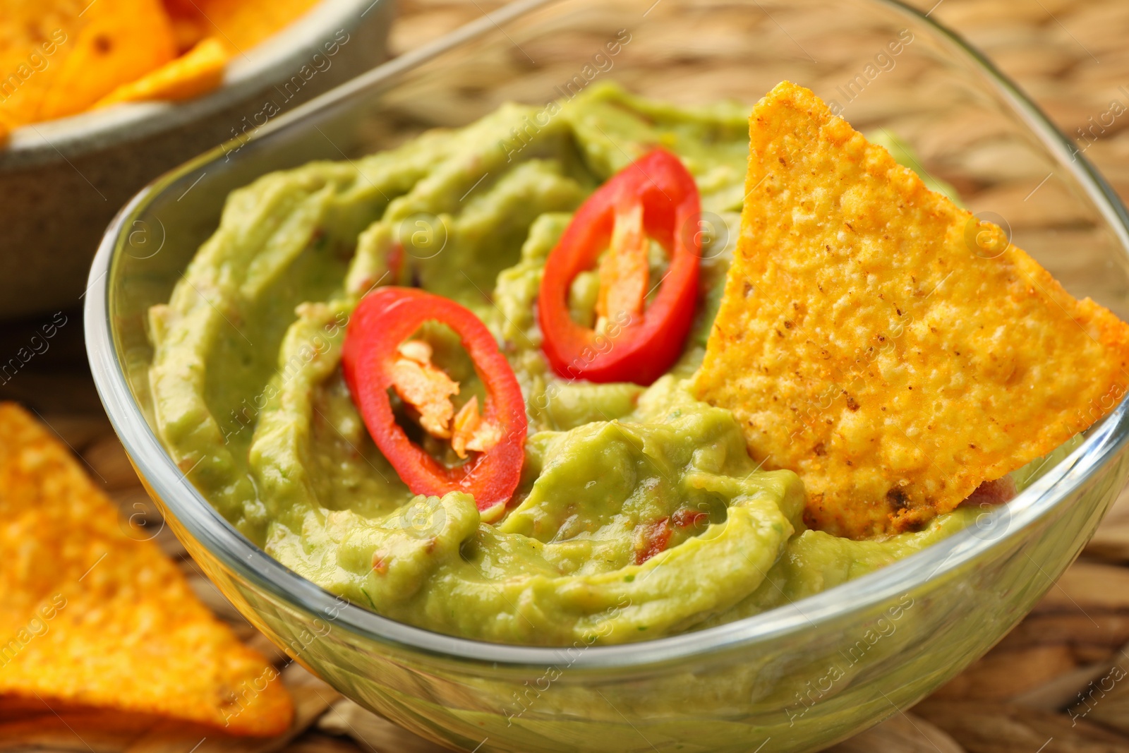 Photo of Bowl of delicious guacamole with chili pepper and nachos chips on wicker table, closeup