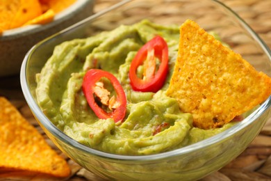 Bowl of delicious guacamole with chili pepper and nachos chips on wicker table, closeup