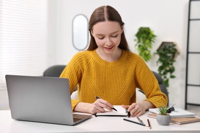 Photo of E-learning. Young woman taking notes during online lesson at white table indoors