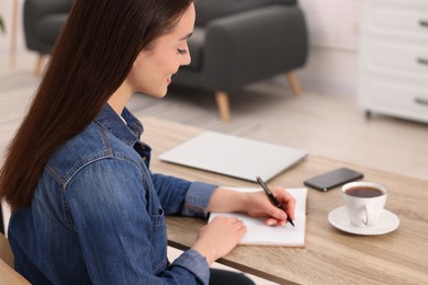 Photo of Young woman writing in notebook at wooden table indoors