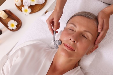 Photo of Woman receiving facial massage with metal roller in beauty salon, top view