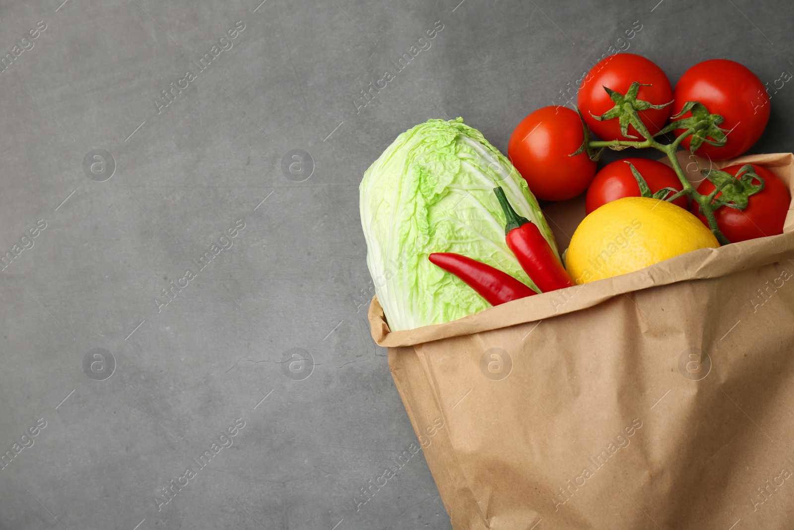 Photo of Paper bag with fresh Chinese cabbage, lemon, tomatoes and chili pepper on grey textured table, top view. Space for text