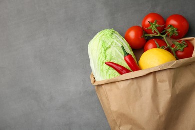 Paper bag with fresh Chinese cabbage, lemon, tomatoes and chili pepper on grey textured table, top view. Space for text