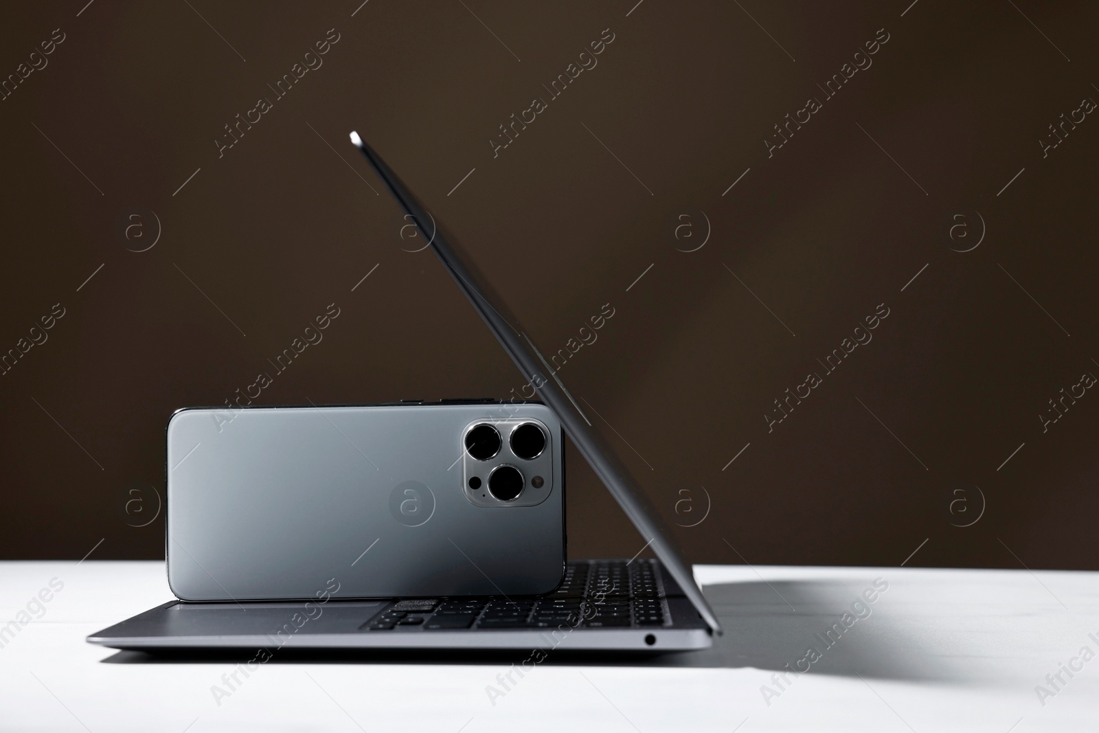 Photo of Modern laptop and smartphones on white table against brown background. Space for text