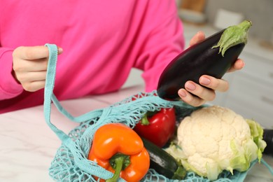 Photo of Woman taking eggplant out from string bag at light marble table, closeup