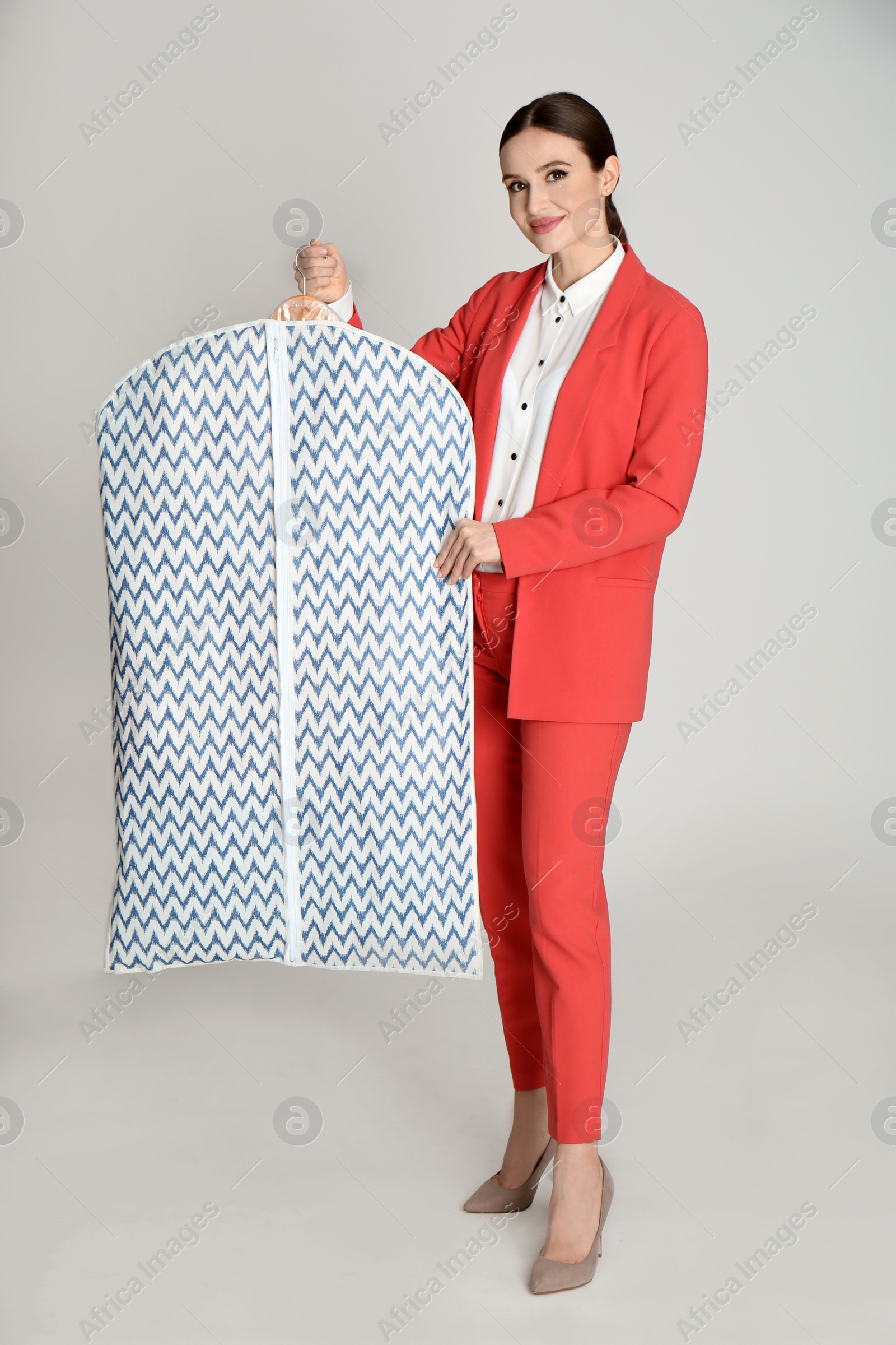 Photo of Young woman holding hanger with clothes in garment cover on light grey background. Dry-cleaning service