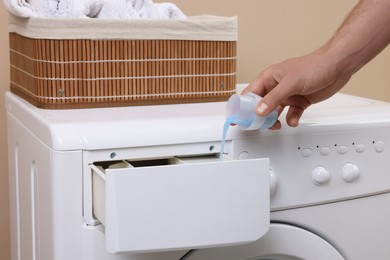 Photo of Man pouring fabric softener from cap into washing machine indoors, closeup