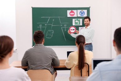 Photo of Teacher showing No Overtaking road sign to audience near chalkboard in driving school