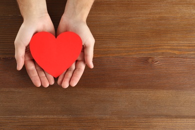 Man holding paper heart on wooden background, top view with space for text