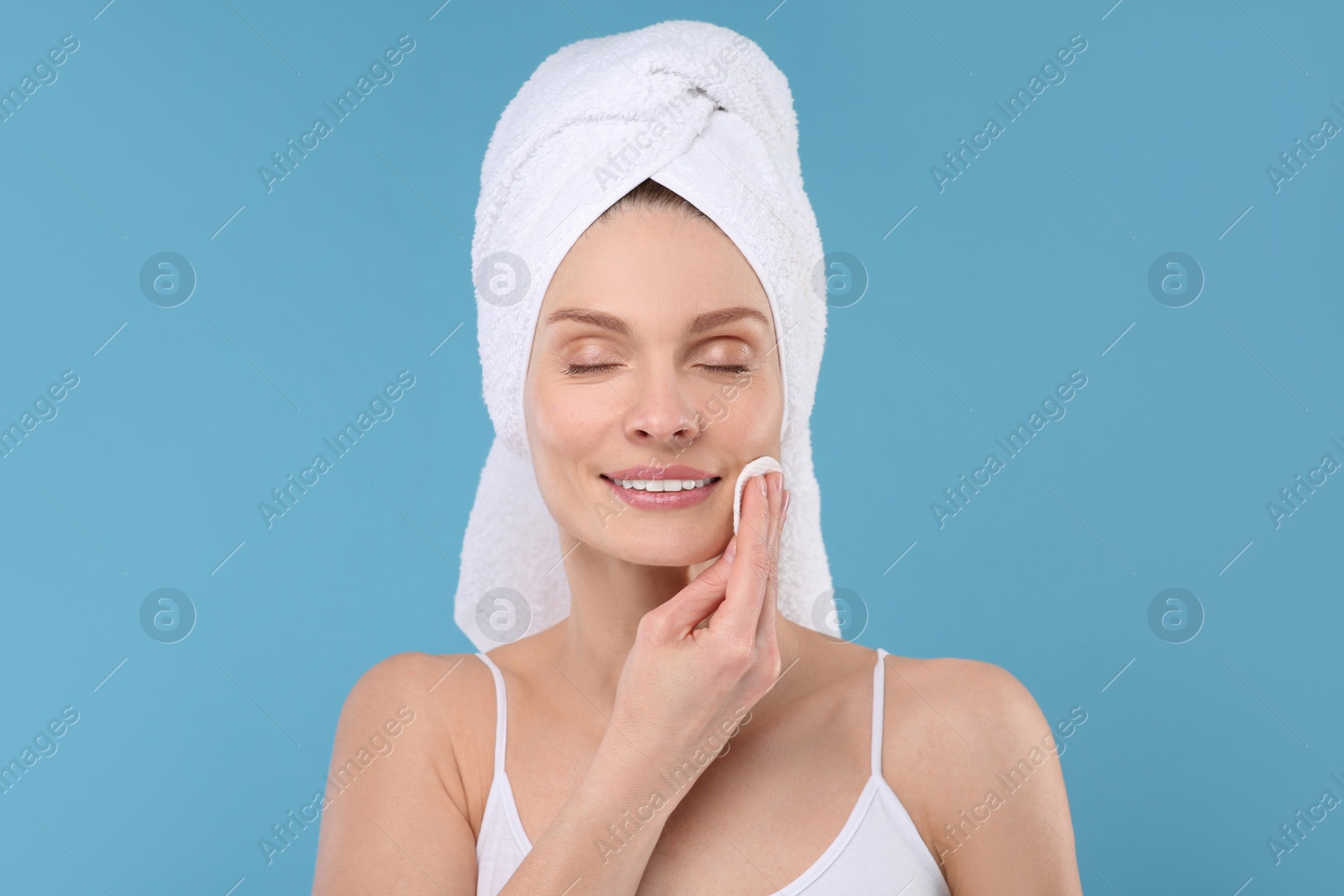 Photo of Beautiful woman in terry towel removing makeup with cotton pad on light blue background