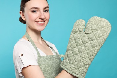 Beautiful young woman in clean apron with pattern and oven glove on light blue background, closeup