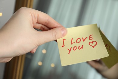 Photo of Woman holding sticky note with phrase I Love You near mirror, closeup