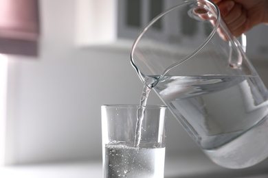Photo of Woman pouring water from jug into glass in kitchen, closeup