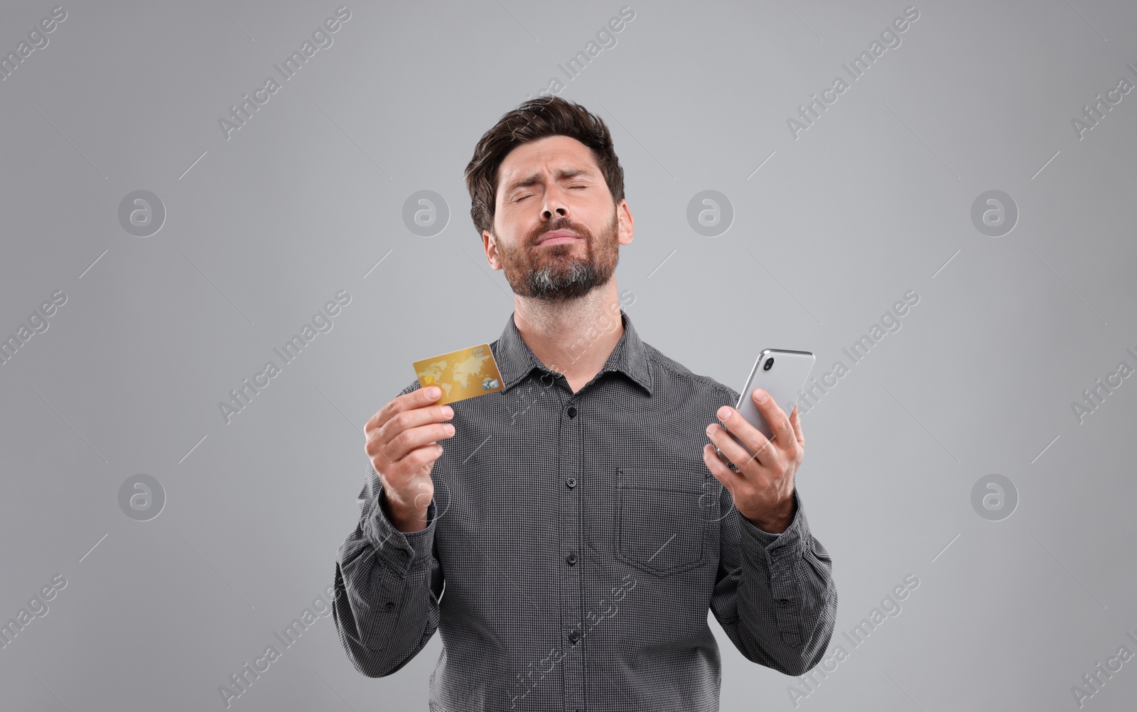 Photo of Upset man with smartphone and credit card on light grey background. Be careful - fraud