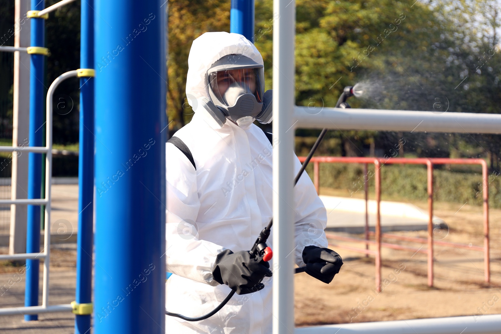 Photo of Man in hazmat suit spraying disinfectant on outdoor gym's equipment. Surface treatment during coronavirus pandemic