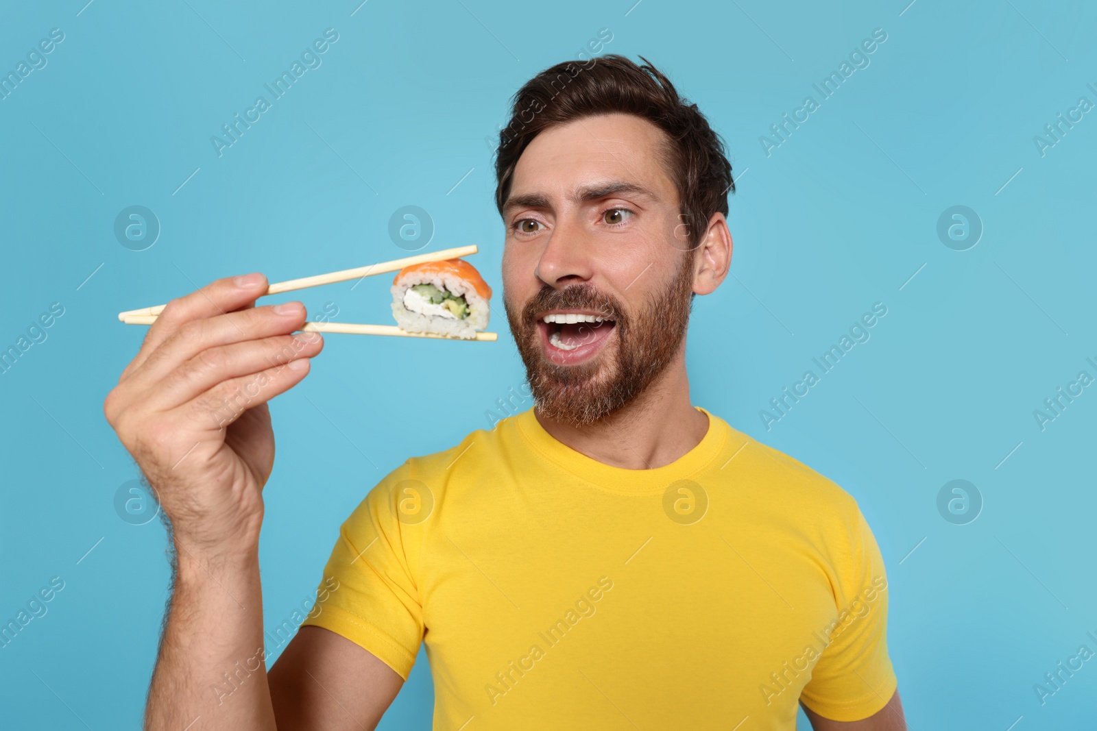 Photo of Happy man eating sushi roll with chopsticks on light blue background