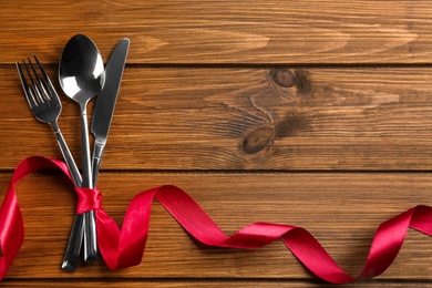 Photo of Cutlery set and red ribbon on wooden background, flat lay with space for text. Valentine's Day dinner