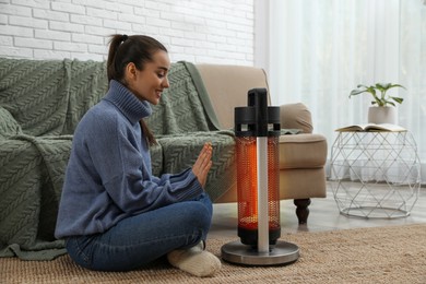 Young woman warming hands near electric heater at home