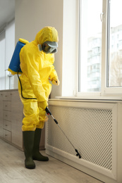 Photo of Pest control worker in protective suit spraying pesticide near window indoors