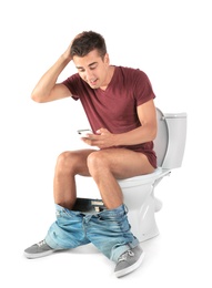 Young man using mobile phone while sitting on toilet bowl. Isolated on white