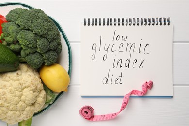 Notebook with words Low Glycemic Index Diet, measuring tape and healthy products on white wooden table, flat lay