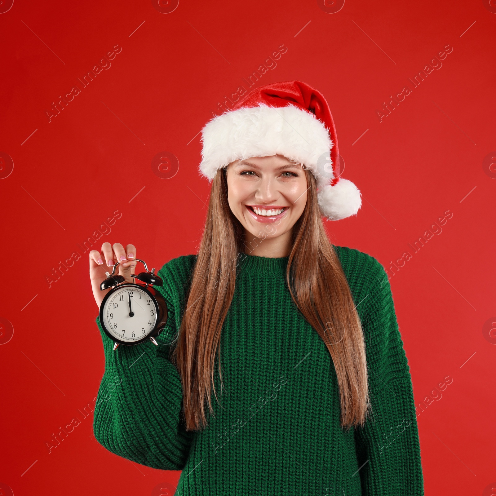 Photo of Happy  young woman in Santa hat with alarm clock on red background. Christmas time