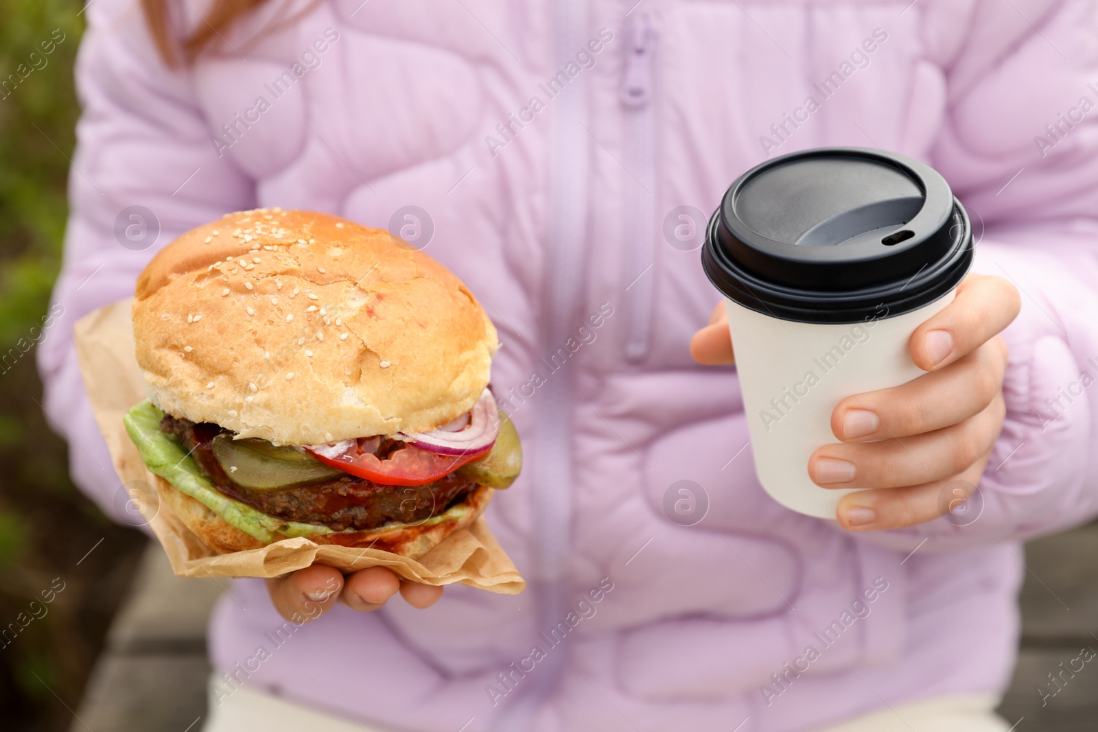 Photo of Little girl holding fresh delicious burger and cup of coffee outdoors, closeup. Street food