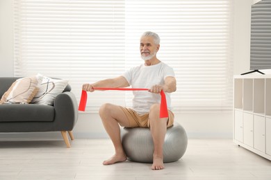 Photo of Senior man doing exercise with elastic resistance band on fitness ball at home