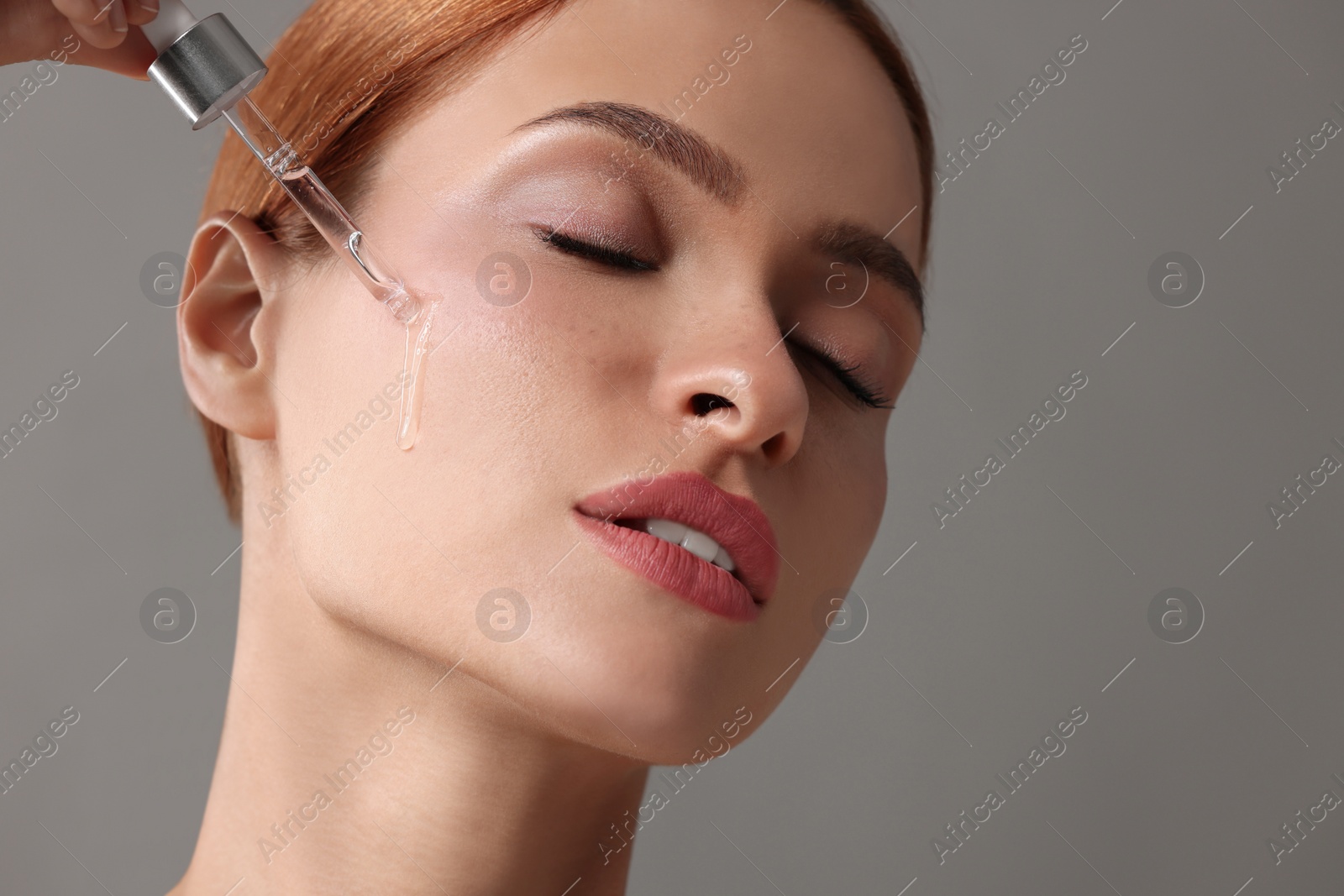 Photo of Beautiful young woman applying cosmetic serum onto her face on grey background, space for text