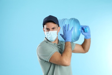 Photo of Courier in face mask with bottle of cooler water on light blue background. Delivery during coronavirus quarantine