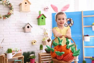 Photo of Adorable little girl with bunny ears and basket full of toy carrots in Easter photo zone