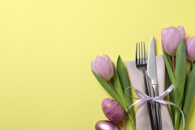 Photo of Cutlery set, Easter eggs and tulips on pale yellow background, flat lay with space for text. Festive table setting