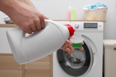 Photo of Man pouring fabric softener from bottle into cap near washing machine indoors, closeup