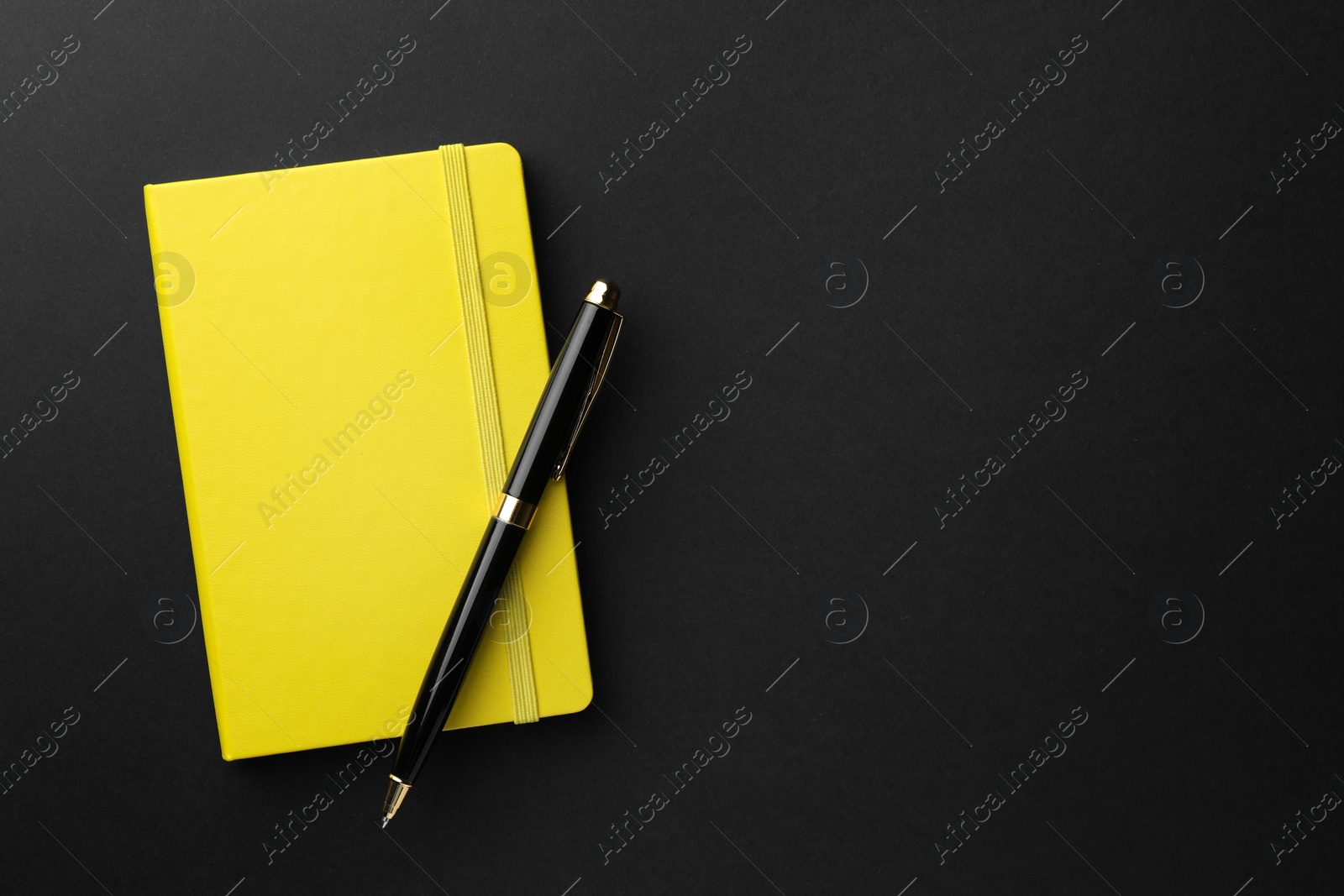 Photo of Closed yellow notebook and pen on black background, top view. Space for text