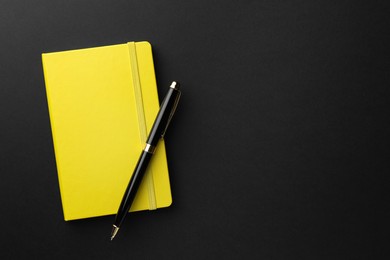 Photo of Closed yellow notebook and pen on black background, top view. Space for text