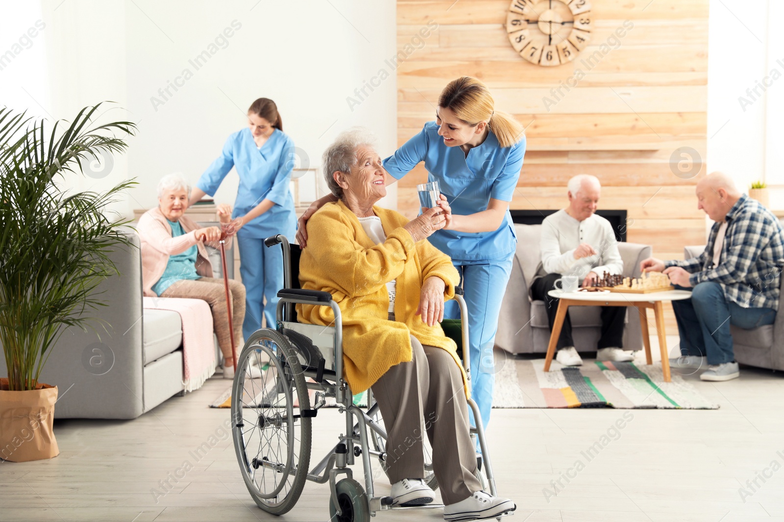 Photo of Nurse giving glass of water to elderly woman in wheelchair at retirement home. Assisting senior people