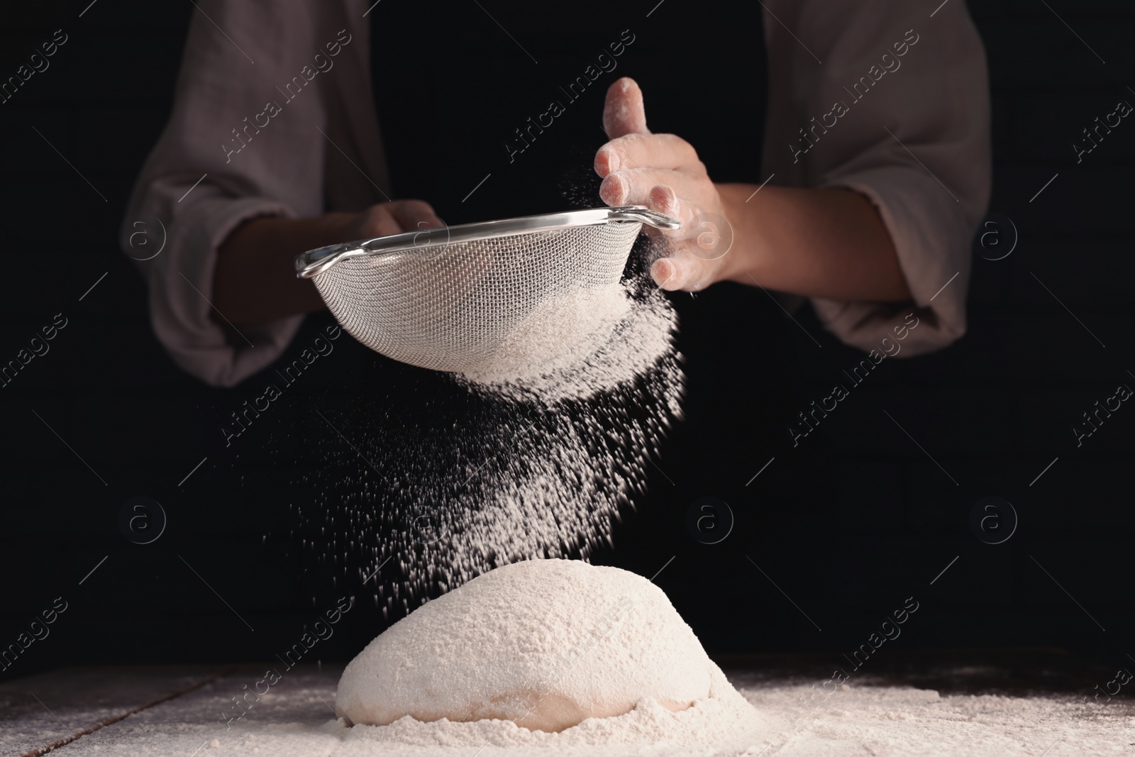 Photo of Woman sprinkling flour over dough at wooden table on dark background, closeup