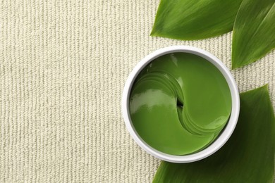 Jar of under eye patches and green leaves on light cloth, top view with space for text. Cosmetic product