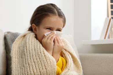 Girl blowing nose in tissue on sofa in room. Cold symptoms