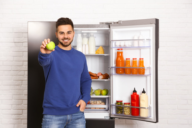 Photo of Happy young man with apple near open refrigerator indoors