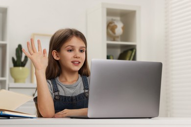 Cute girl waving hello during online lesson via laptop at white table indoors