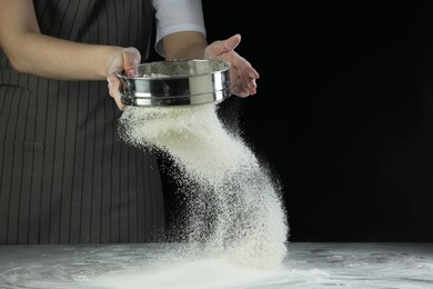 Woman sieving flour at table against black background, closeup. Space for text