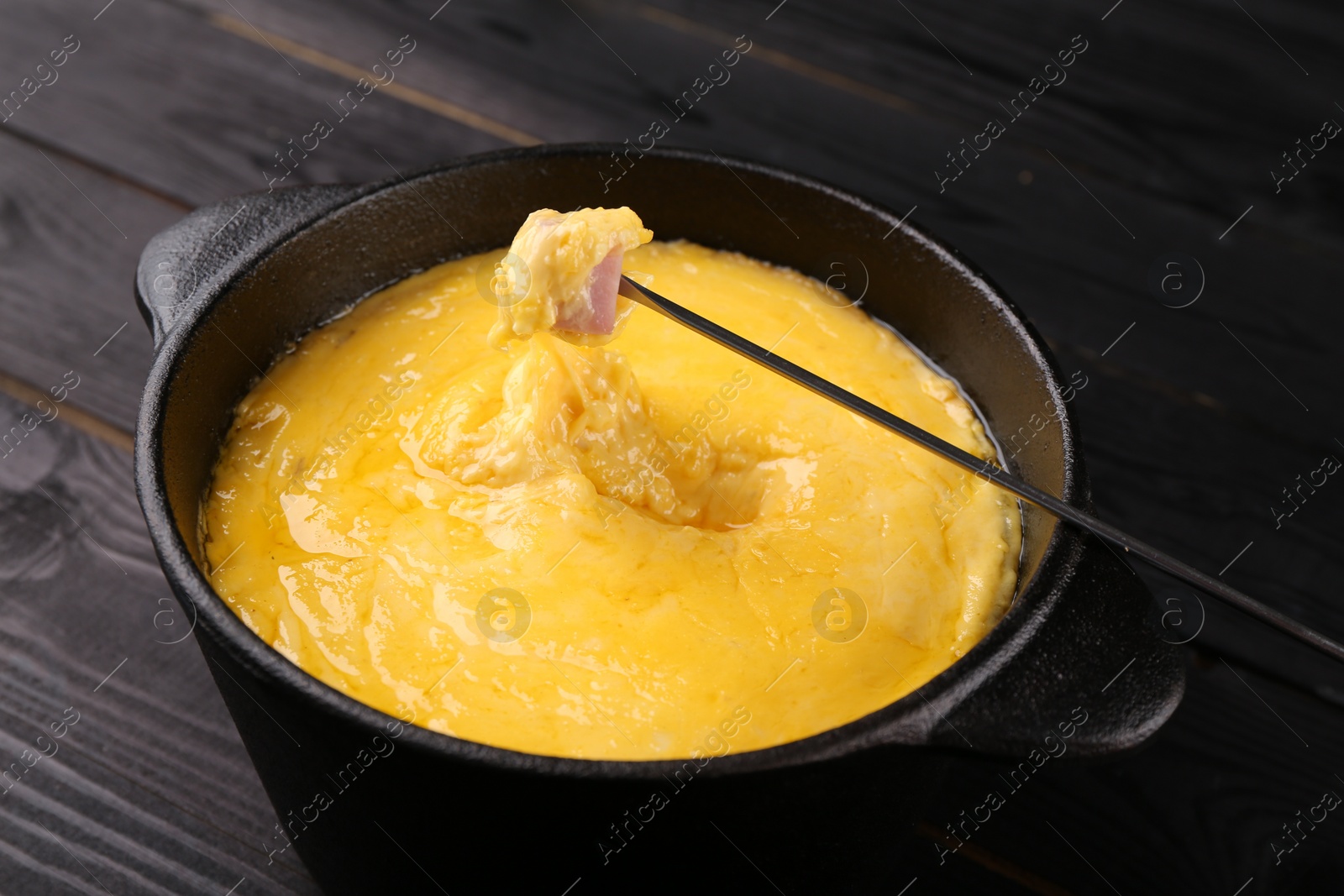 Photo of Dipping piece of ham into fondue pot with melted cheese on black wooden table, closeup