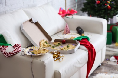 Photo of Paper plate with piece of pizza on messy sofa indoors. Chaos after party